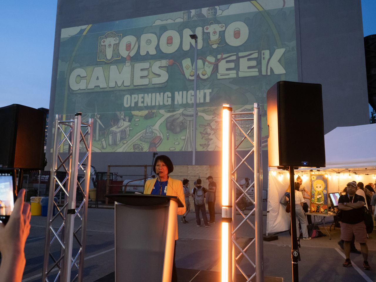 Mayor Olivia Chow speaks at a podium in front of the waterfront malting silos, on which "Toronto Games Week" is projected.