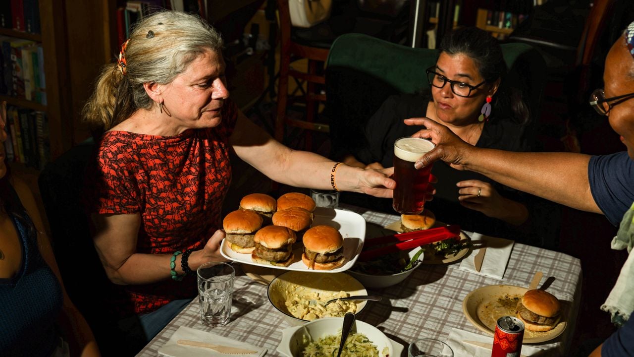 A person at a table holds a tray of burgers while another person passes them a pint of beer