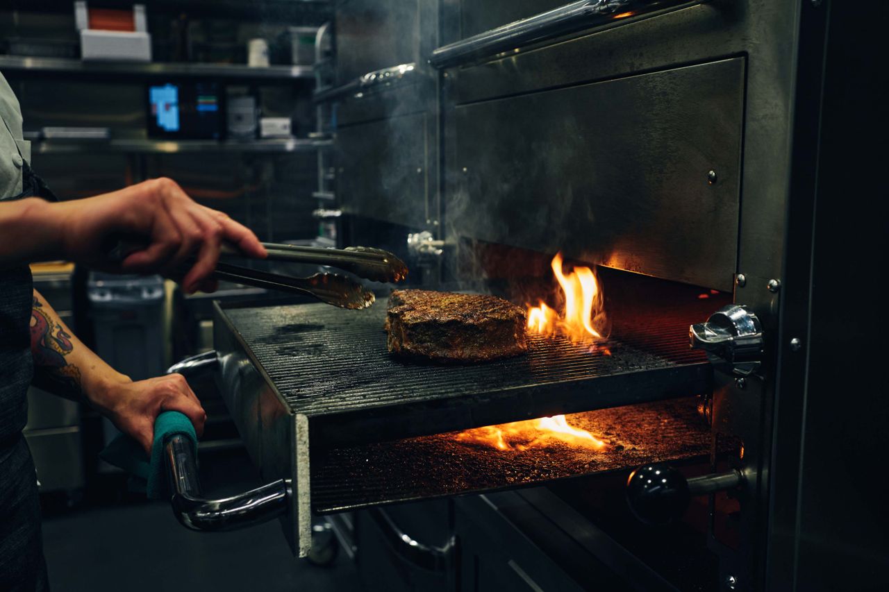 A chef holding tongs grills a steak over an open flame in the kitchen of a restaurant