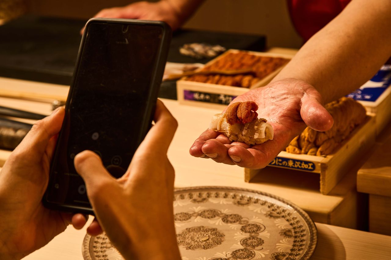 A chef hands a piece of sushi to a customer, who takes a photo using their phone
