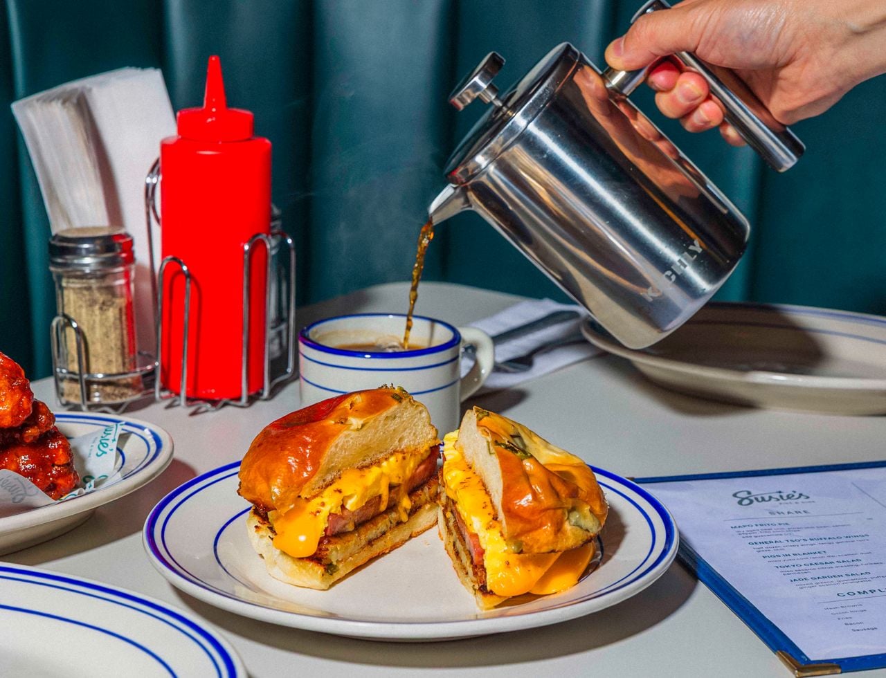 A person pours coffee into a mug placed behind a plate with a breakfast sandwich on it