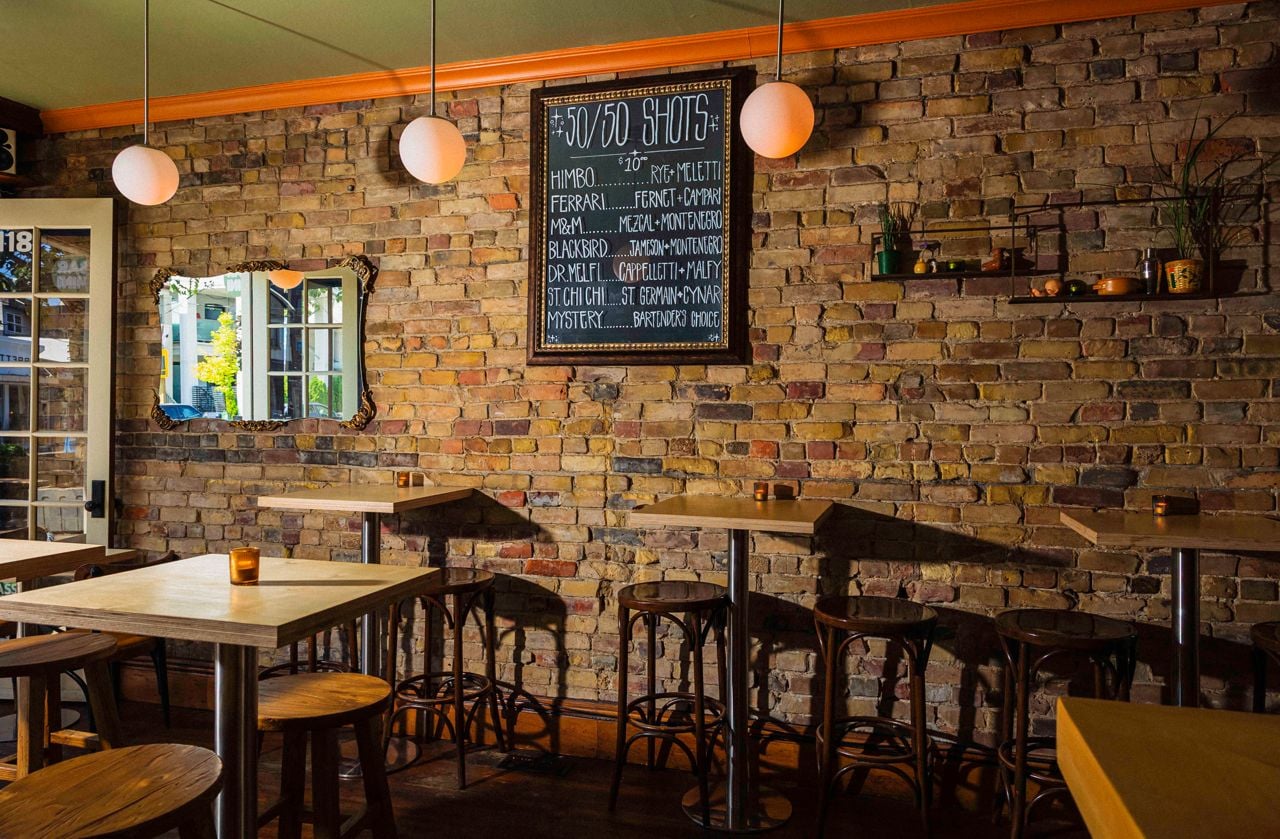 The dining room at Bar Mini, with one wall of exposed brick and a chalkboard menu listing shots