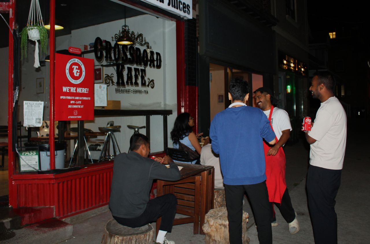 People sit at tables on the sidewalk outside of Crossroad Kafe and eat burgers and fried chicken from Those Guys