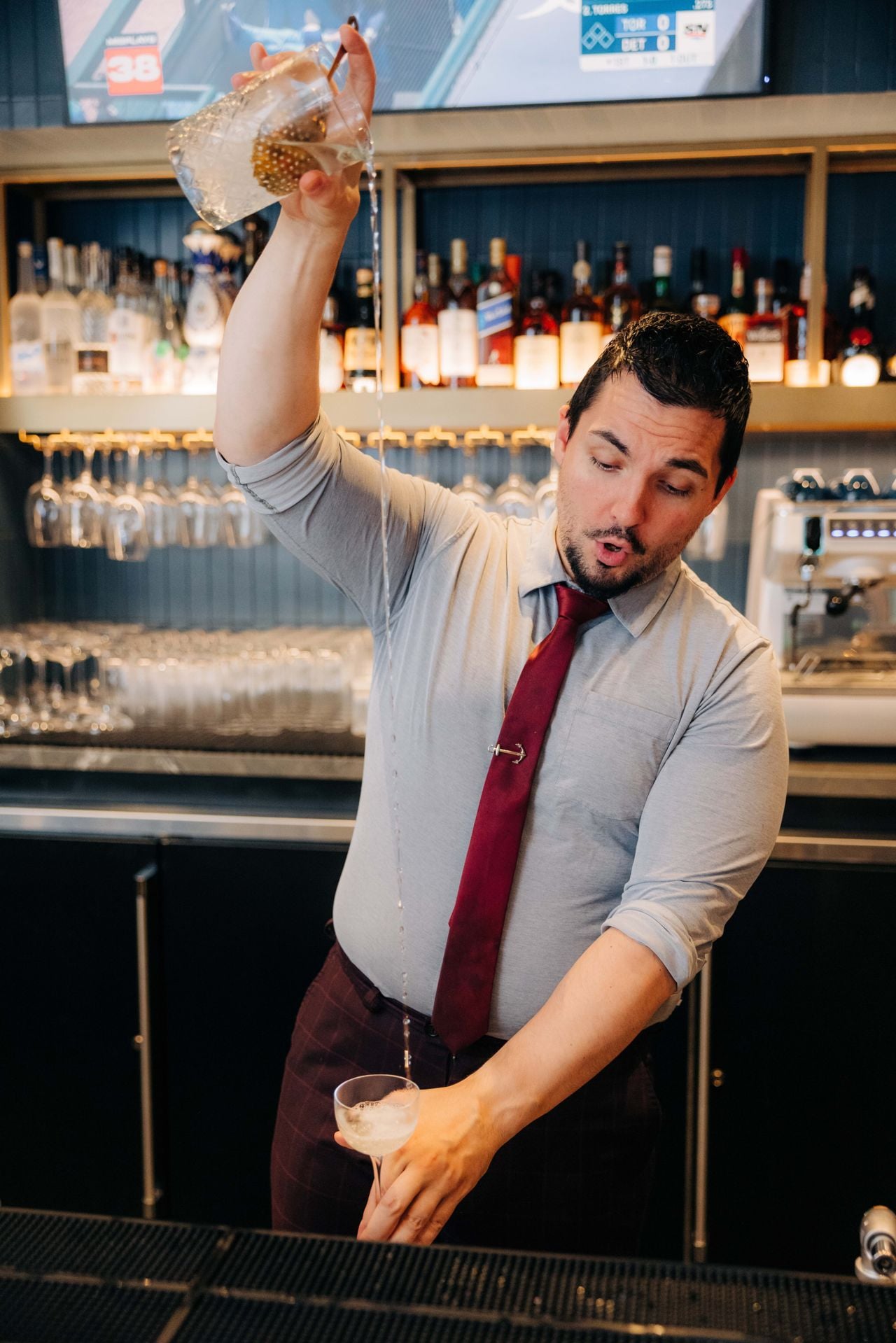 A bartender pours a drink from a mixing glass into a coupe from a great height
