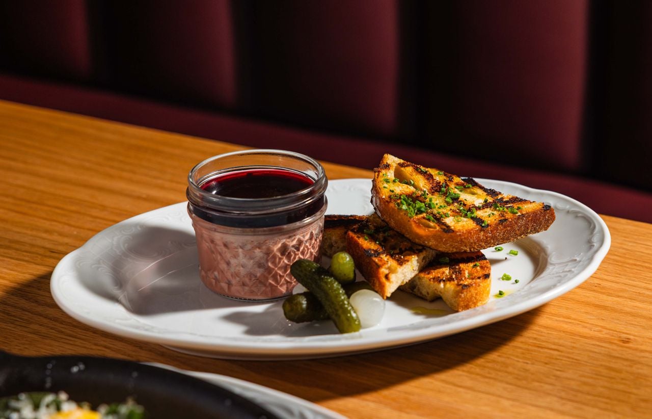 Chicken liver pâté is topped with a reduced red wine jelly and paired with bread-and-butter pickles and a pearl onion, and served with sourdough bread