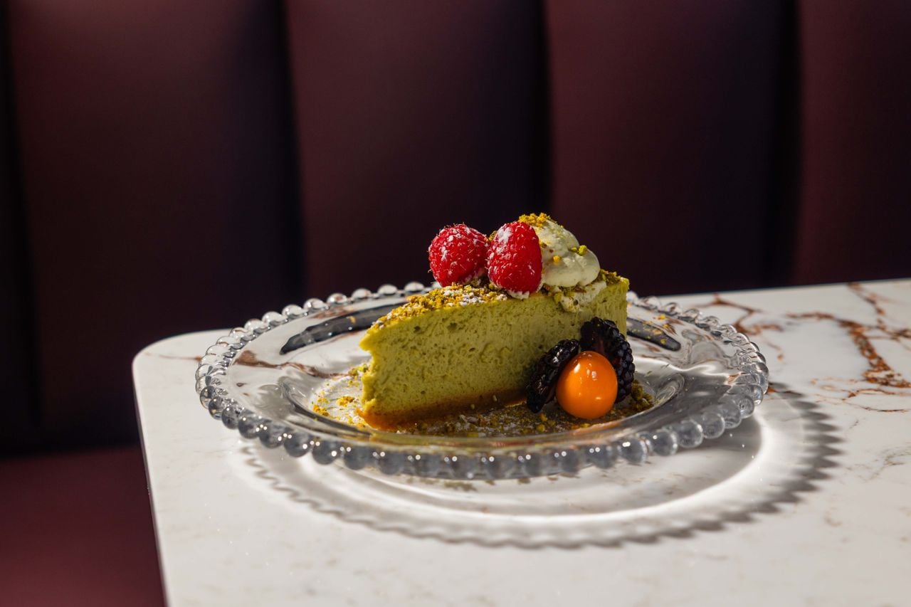A slice of pistachio cheesecake comes topped with pistachio crema, raspberry coulis, crumbled pistachios and fresh berries