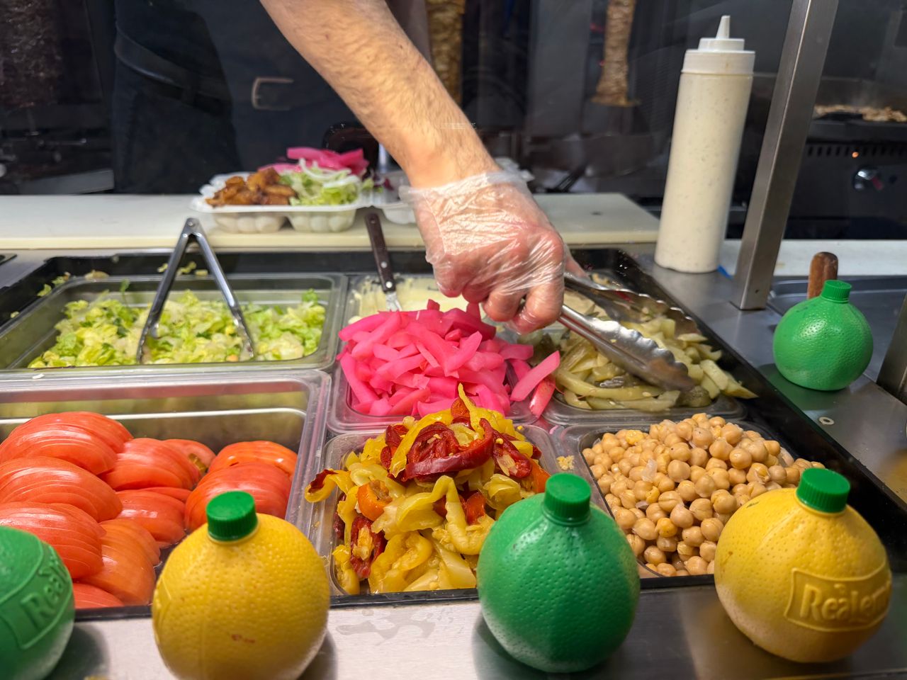 A worker uses tongs to grab pickles from a selection of condiments at a shawarma restaurant