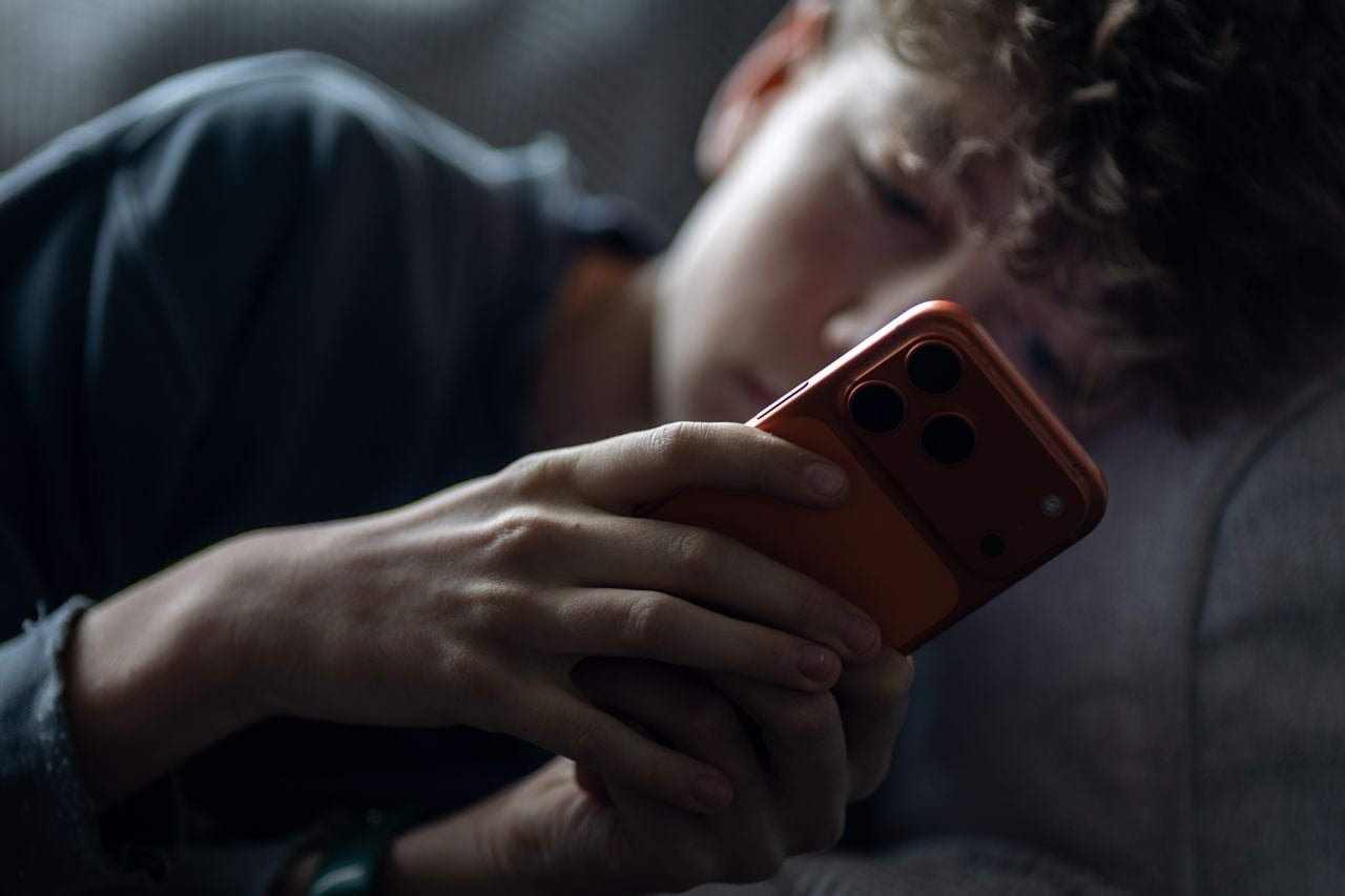A teenaged boy lies in bed looking at his phone.