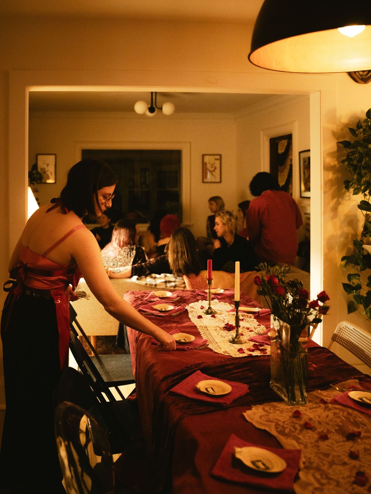 A person sets a dinner table while guests mingle in the background