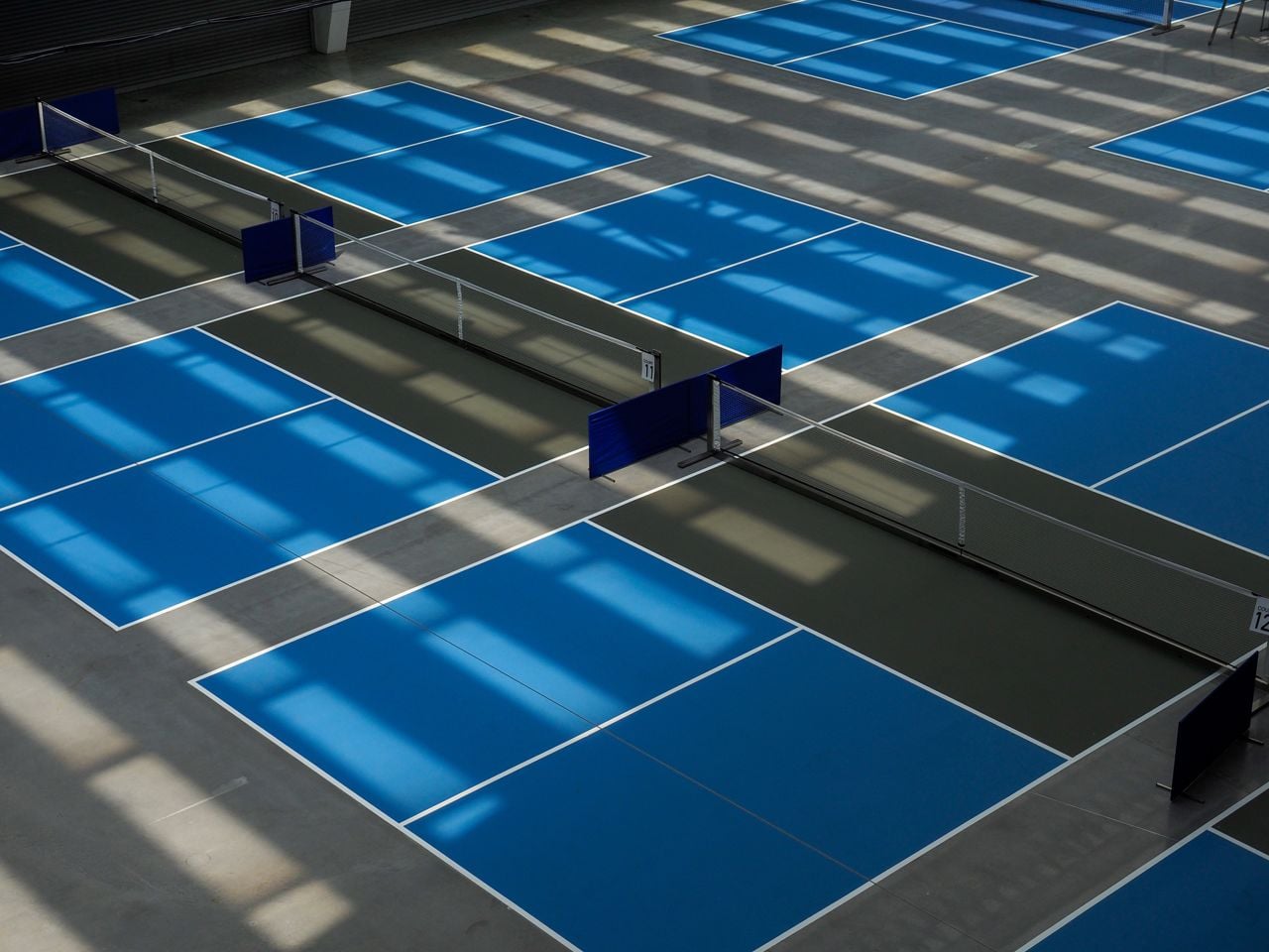 An indoor pickleball court indoor with blue service area and dark grey area known as the kitchen or non volley area with some light and shadows in play.