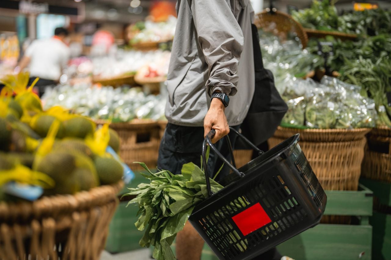 A man walks in the grocery store carrying a basket, which contains a leafy green vegetable.