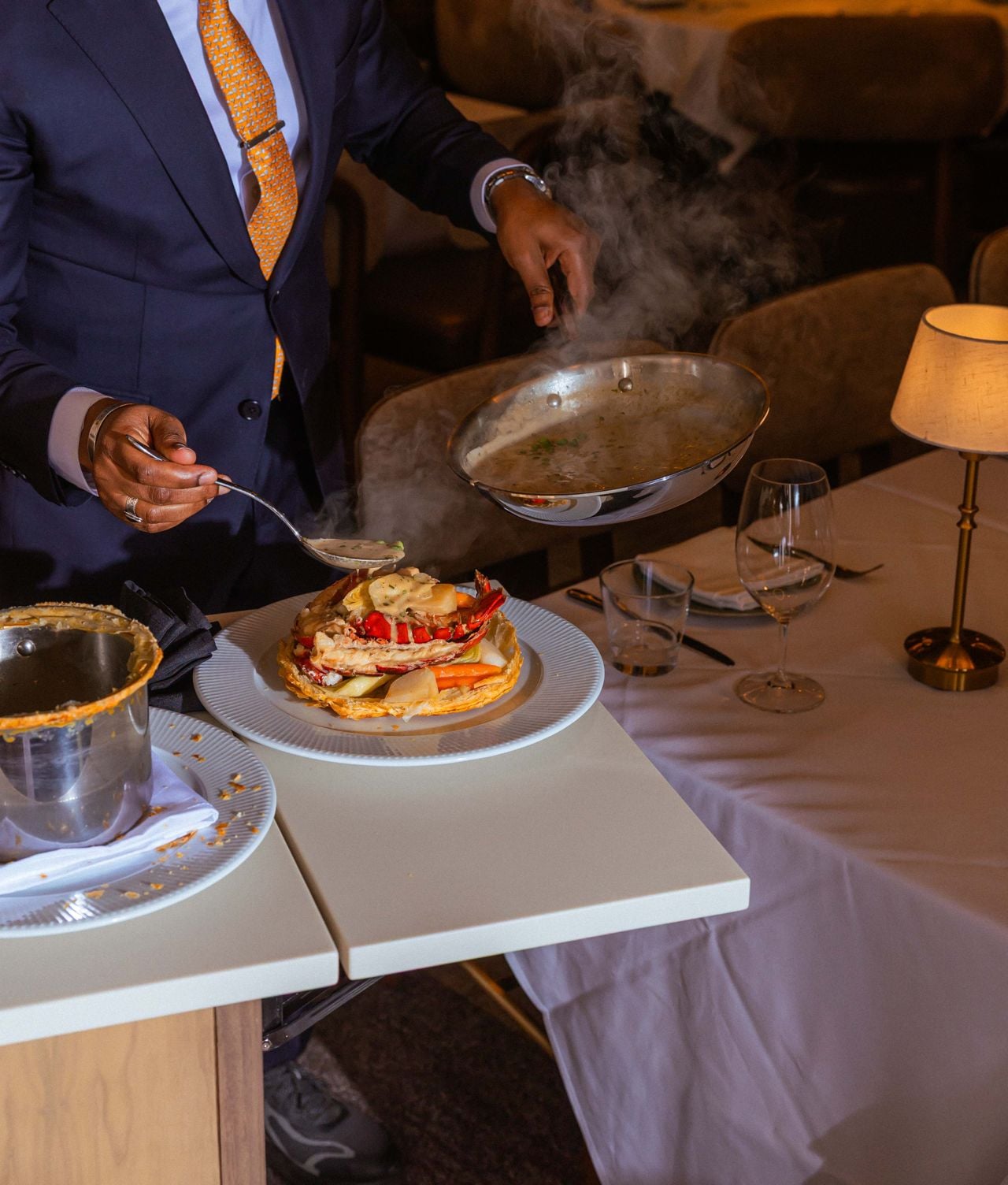 A server pours a spoonful of sauce tableside over a serving of lobster pot pie