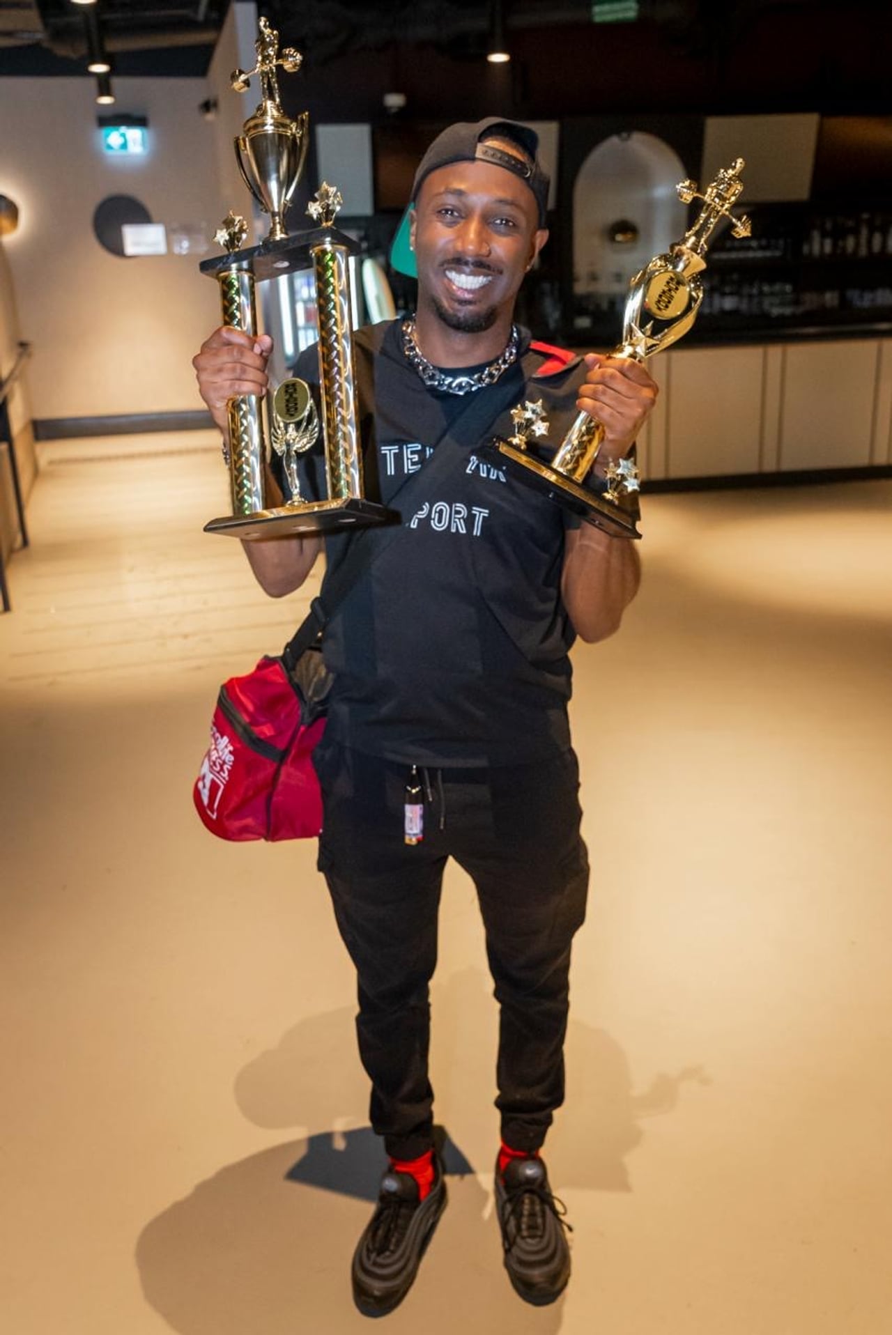 A performer in a black athleisure set with a gym bag and backwards baseball cap holding two trophies