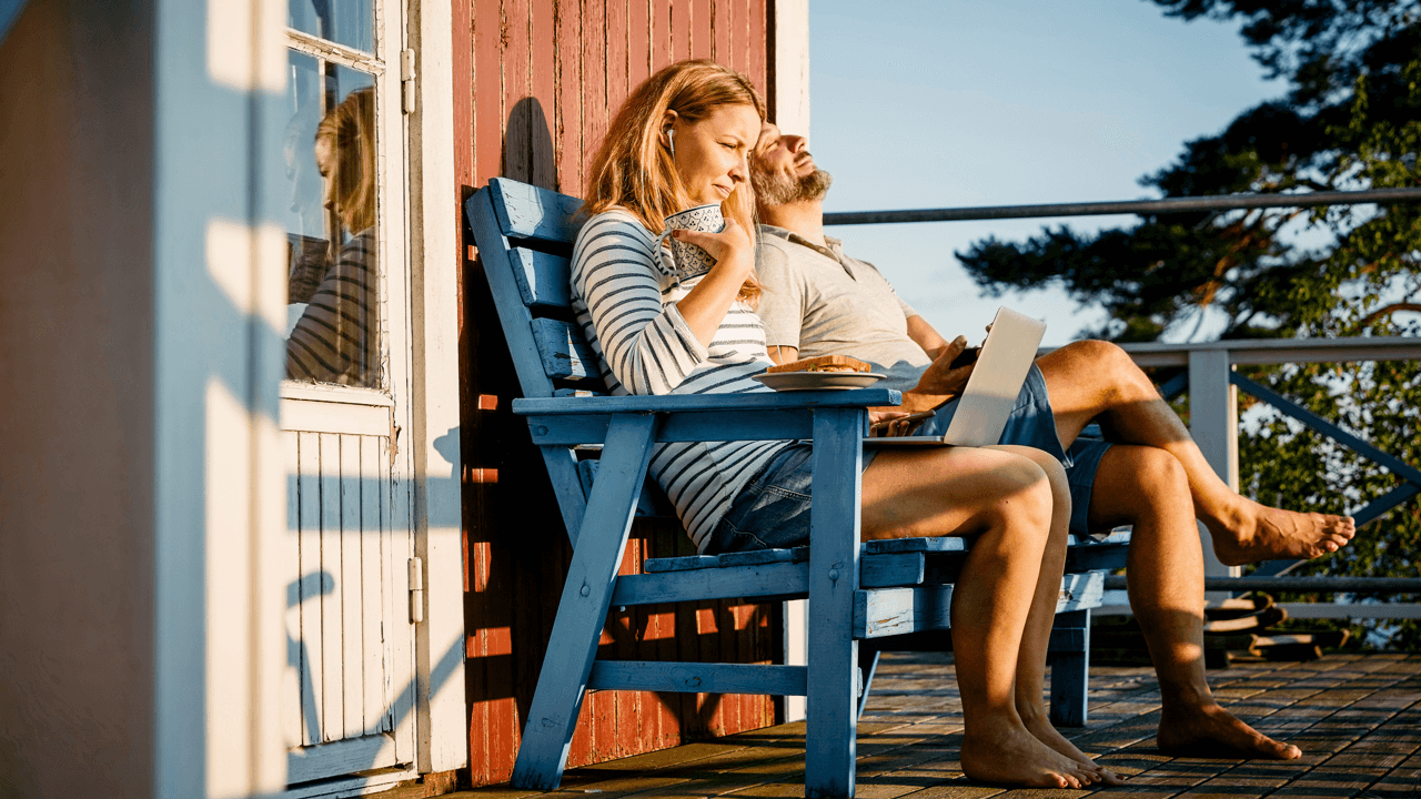 A man and woman sit on the porch of their cottage. The woman looks down at her laptop.