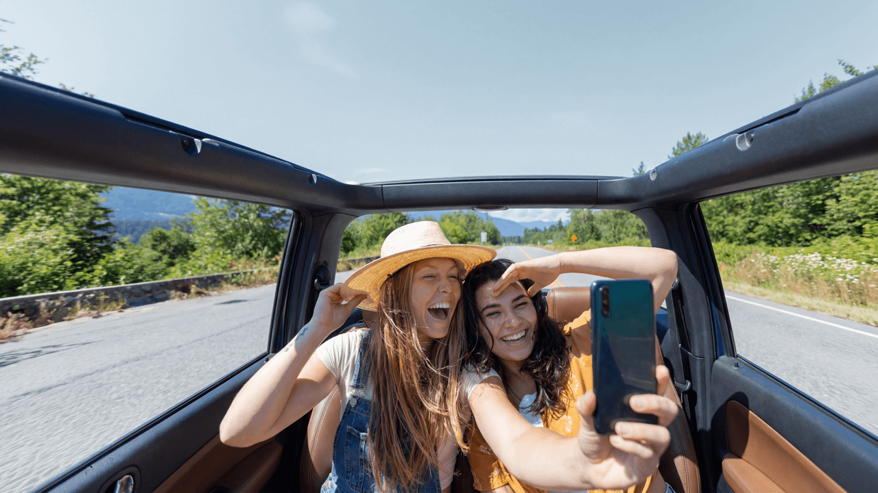 Two girls ride in the back seat of a convertible car and smile as they take a picture on their phone.