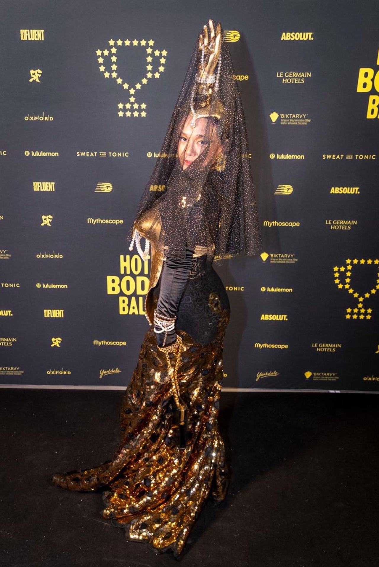 A performer in a black and gold gown with a veil being held up by a gold hand-shaped headdress