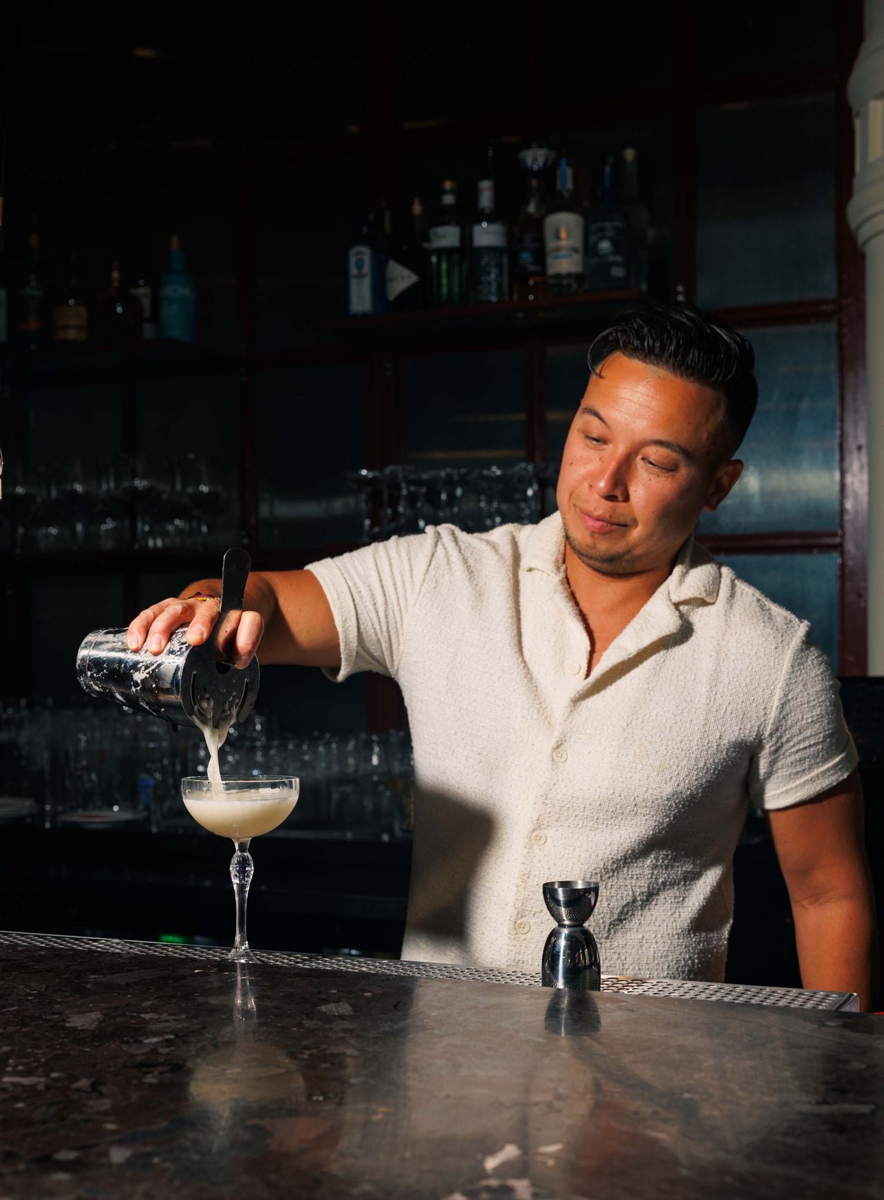 A bartender pours a drink from a shaker into a coupe glass