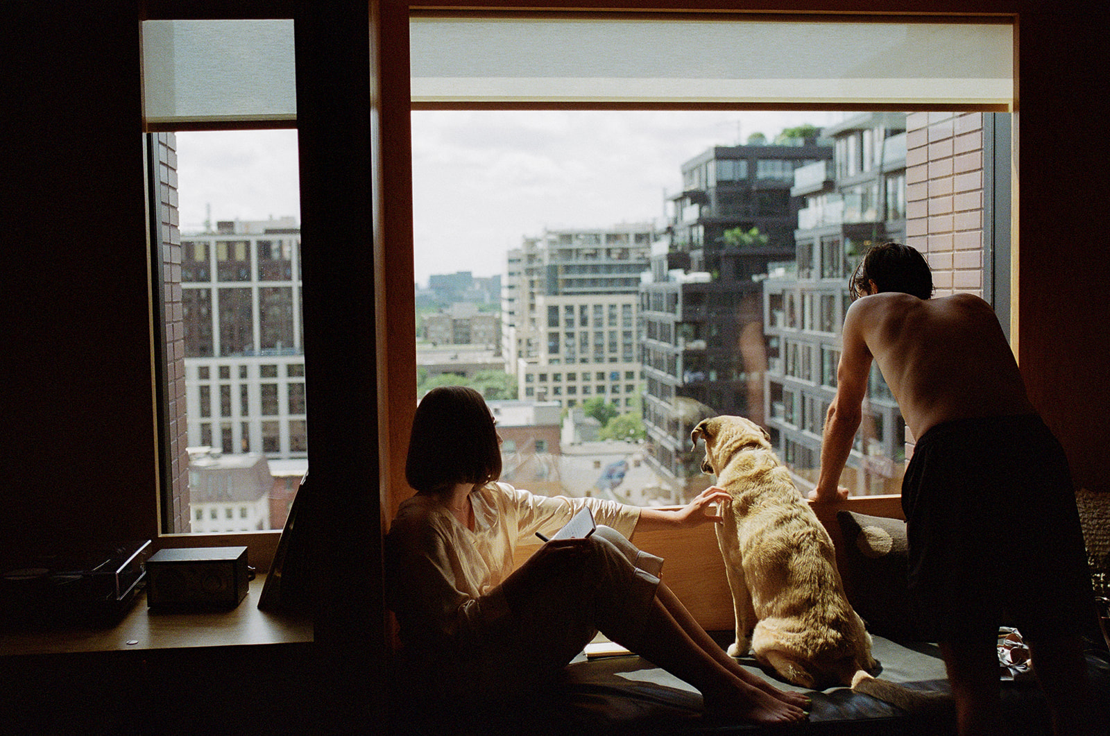 Ryanne, a shirless Brjánn, and their dog look out a window of the Ace Hotel.