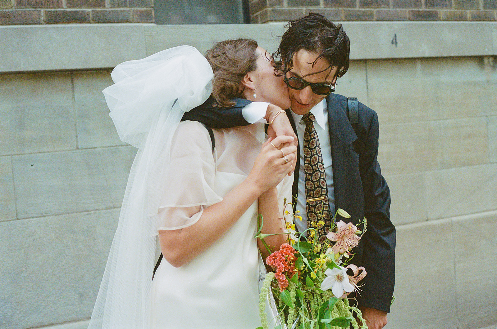 Ryanne kisses Brjánn on the cheek outside a brownstone building.
