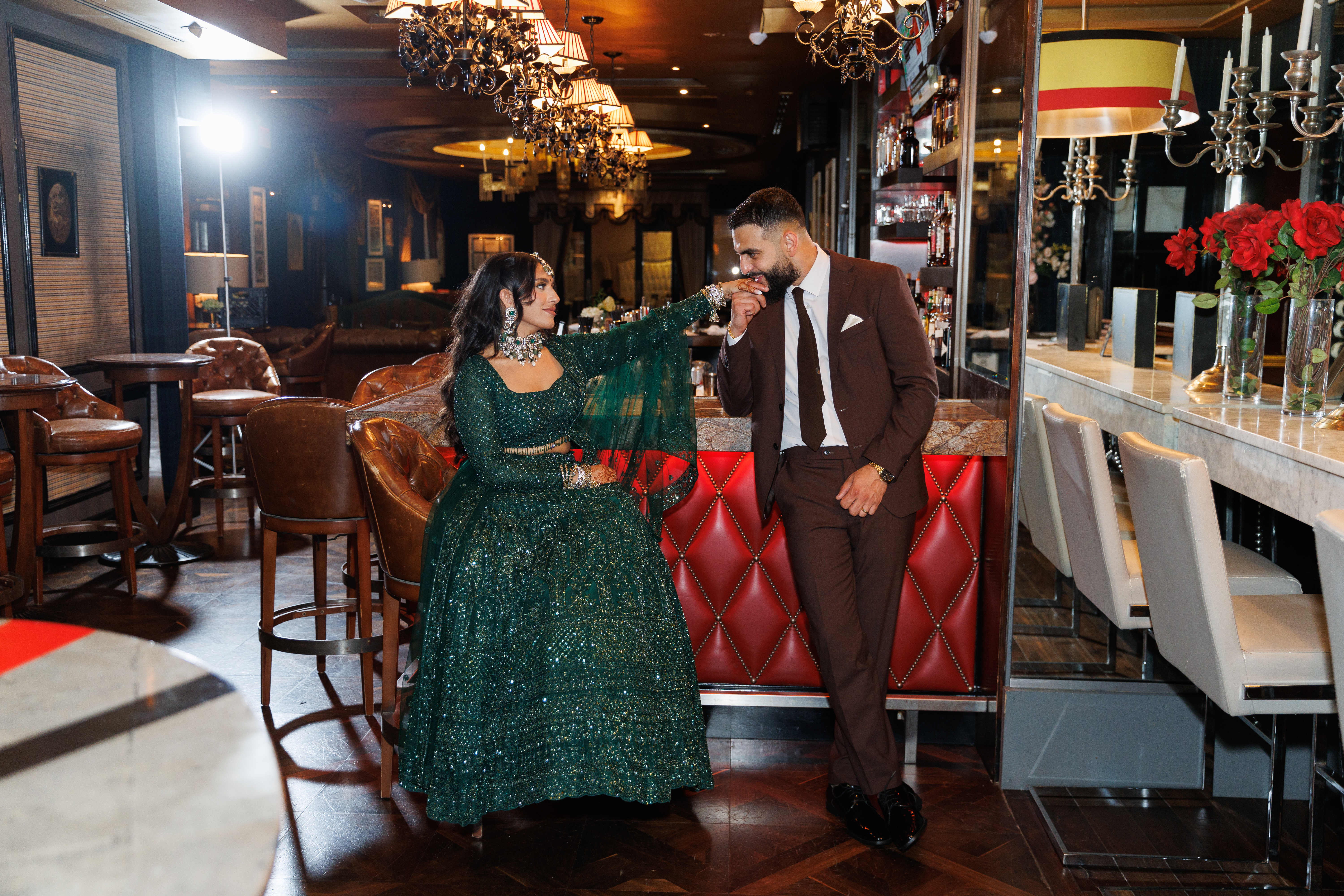 Haris, in a suit and tie, kisses Gital's hand at the bar of the Windsor Arms Hotel. Gital is wearing a sparkling green dress.