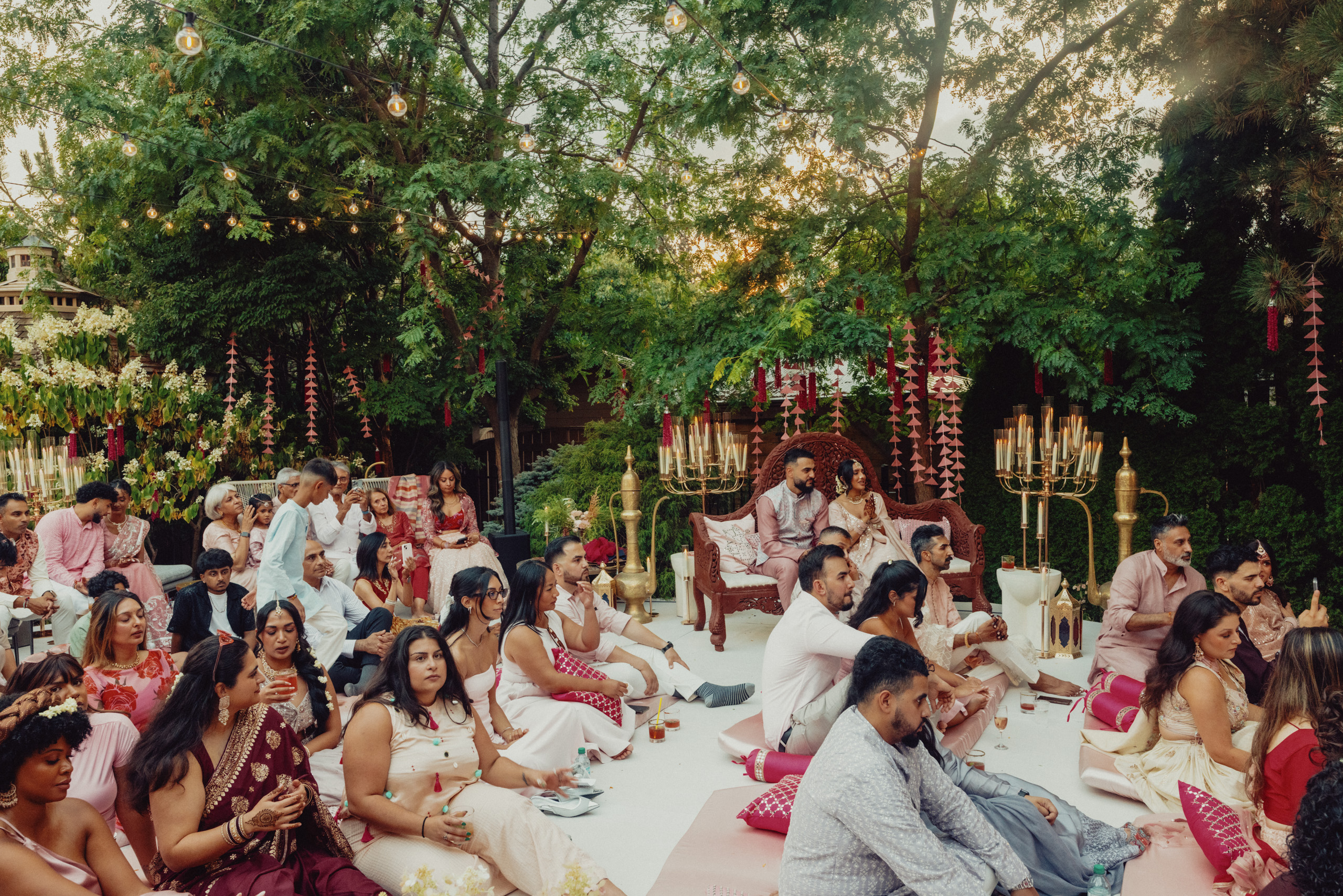 Wedding guests sit cross-legged on a platform at the outdoor mehdi celebration.